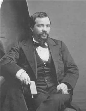 A seated white man with a beard and mustache wearing a suit jacket, vest, and bow tie, holds a small book in his right hand